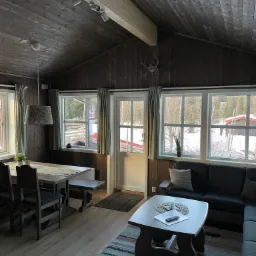 Wnętrze drewnianego domu z widokiem na zimowy krajobraz, drewniane panele na ścianach i suficie, st&oacute;ł z ławką, sofa i stolik kawowy z pilotem i świecą.