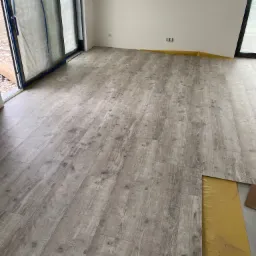 Jasnoszara podłoga laminowana z imitacją drewna, ułożona w nowym pomieszczeniu z zabezpieczonymi folią oknami i listwami przypodłogowymi zabezpieczonymi taśmą malarską, widoczny fragment podkładu.