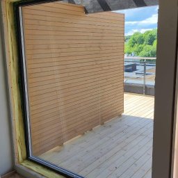 Jakub Parkiety-Malowanie - Montaż okna balkonowego z widokiem na taras z desek w Rawa Mazowiecka, widoczna izolacja wokół ramy okiennej.