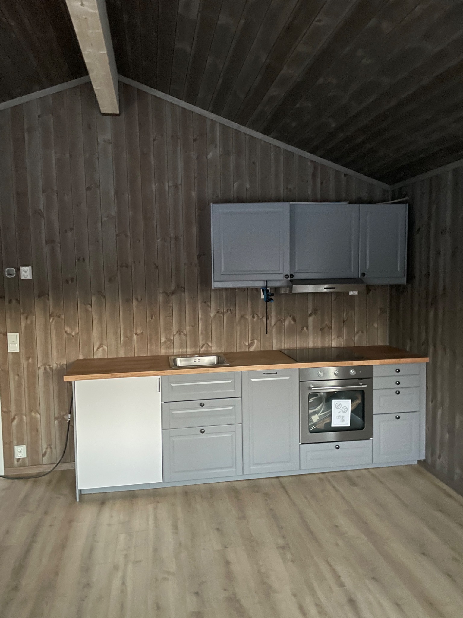Szare szafki kuchenne z drewnianym blatem w pomieszczeniu wykończonym drewnem, widoczny fragment podłogi i sufitu.
