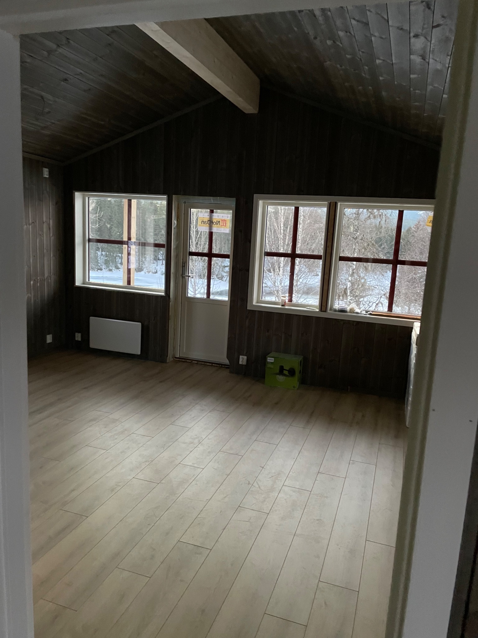 Wnętrze pomieszczenia z drewnianą boazerią na ścianach i suficie, jasną podłogą imitującą drewno, trzema oknami z widokiem na zimowy krajobraz i grzejnikiem elektrycznym pod jednym z nich.