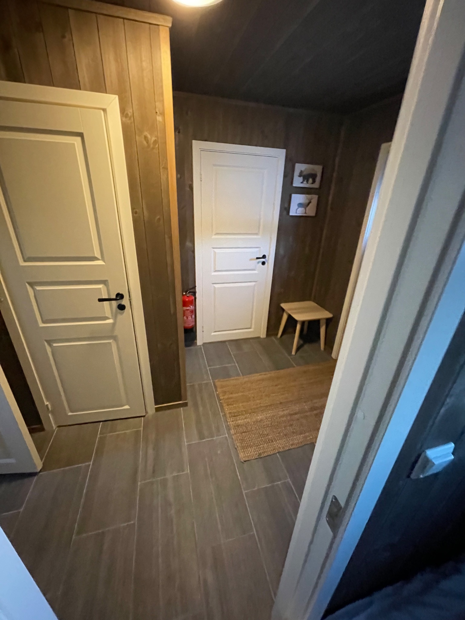 Wnętrze korytarza z drewnianymi ścianami i szarą podłogą z płytek, widoczne białe drzwi z czarnymi klamkami, mały stołek, dywanik i gaśnica.