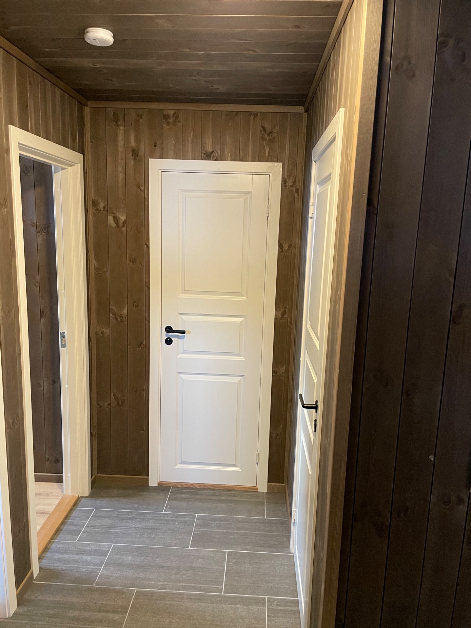 Korytarz wykończony drewnianą boazerią z dwiema białymi, zamkniętymi drzwiami z czarnymi klamkami oraz szarą podłogą z płytek imitujących kamień.