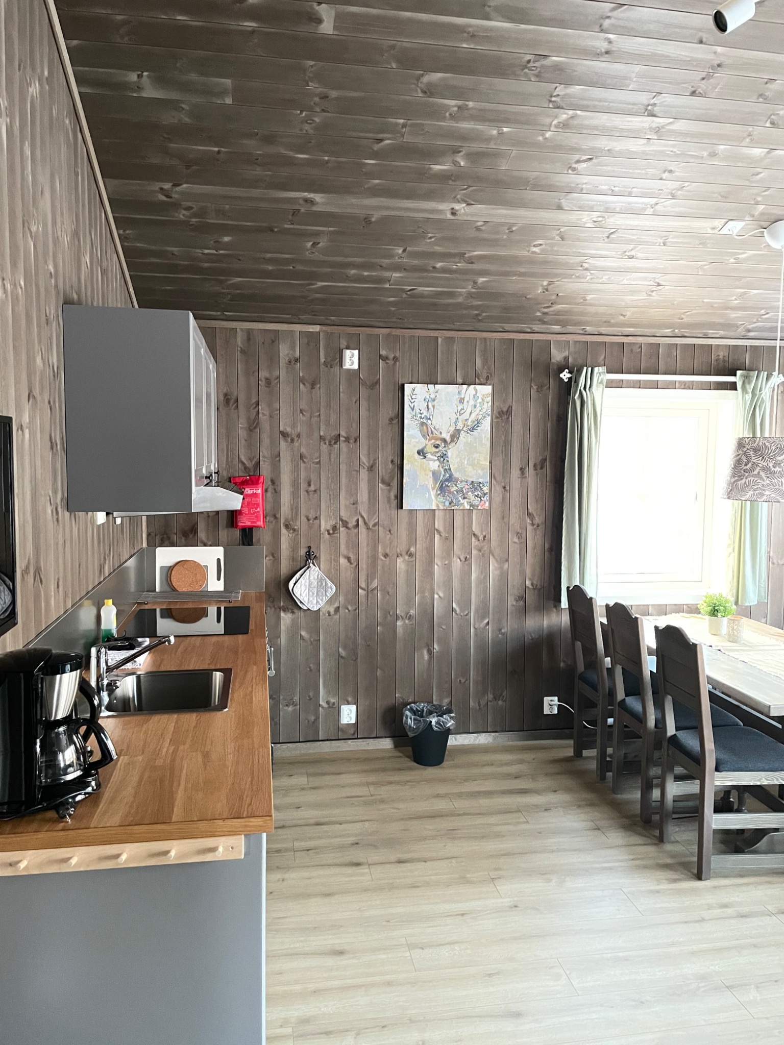Wnętrze kuchni w stylu skandynawskim z drewnianymi panelami ściennymi, szarą zabudową kuchenną z drewnianym blatem, stołem z krzesłami i obrazem jelenia na ścianie.