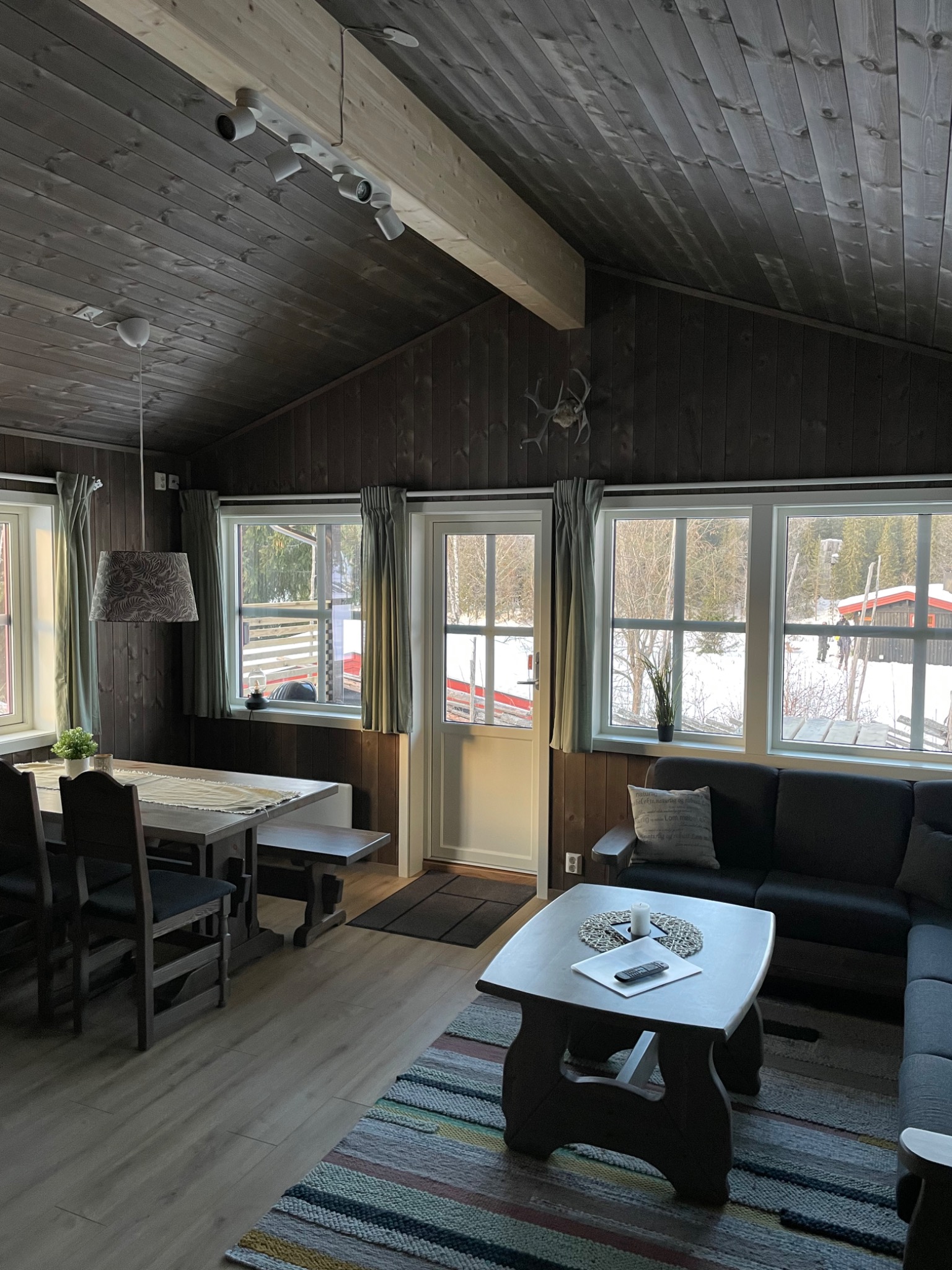 Wnętrze drewnianego domu z widokiem na zimowy krajobraz, drewniane panele na ścianach i suficie, stół z ławką, sofa i stolik kawowy z pilotem i świecą.