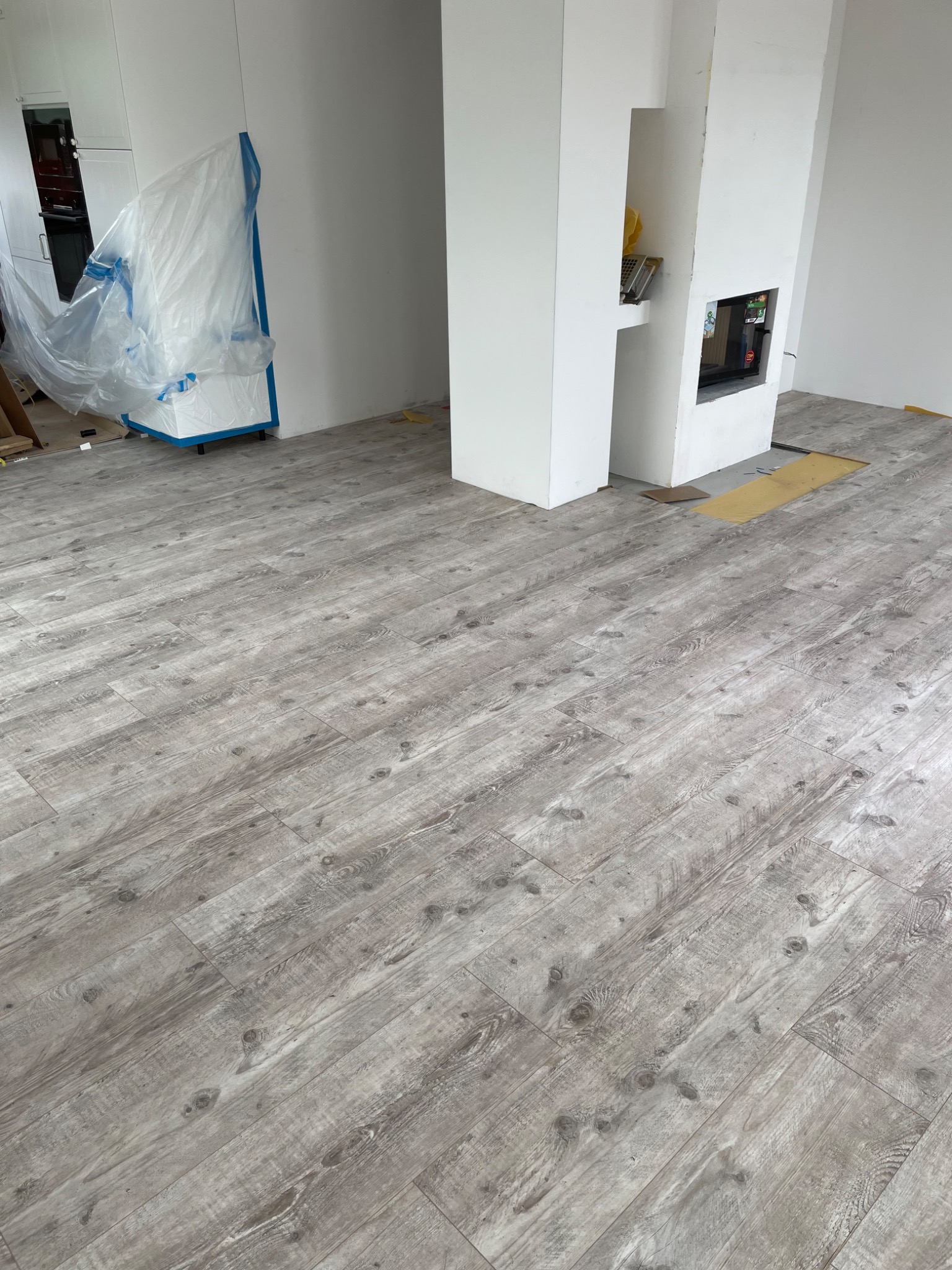 Układanie jasnoszarego parkietu panelowego w pomieszczeniu z kominkiem, zabezpieczone meble folią malarską.