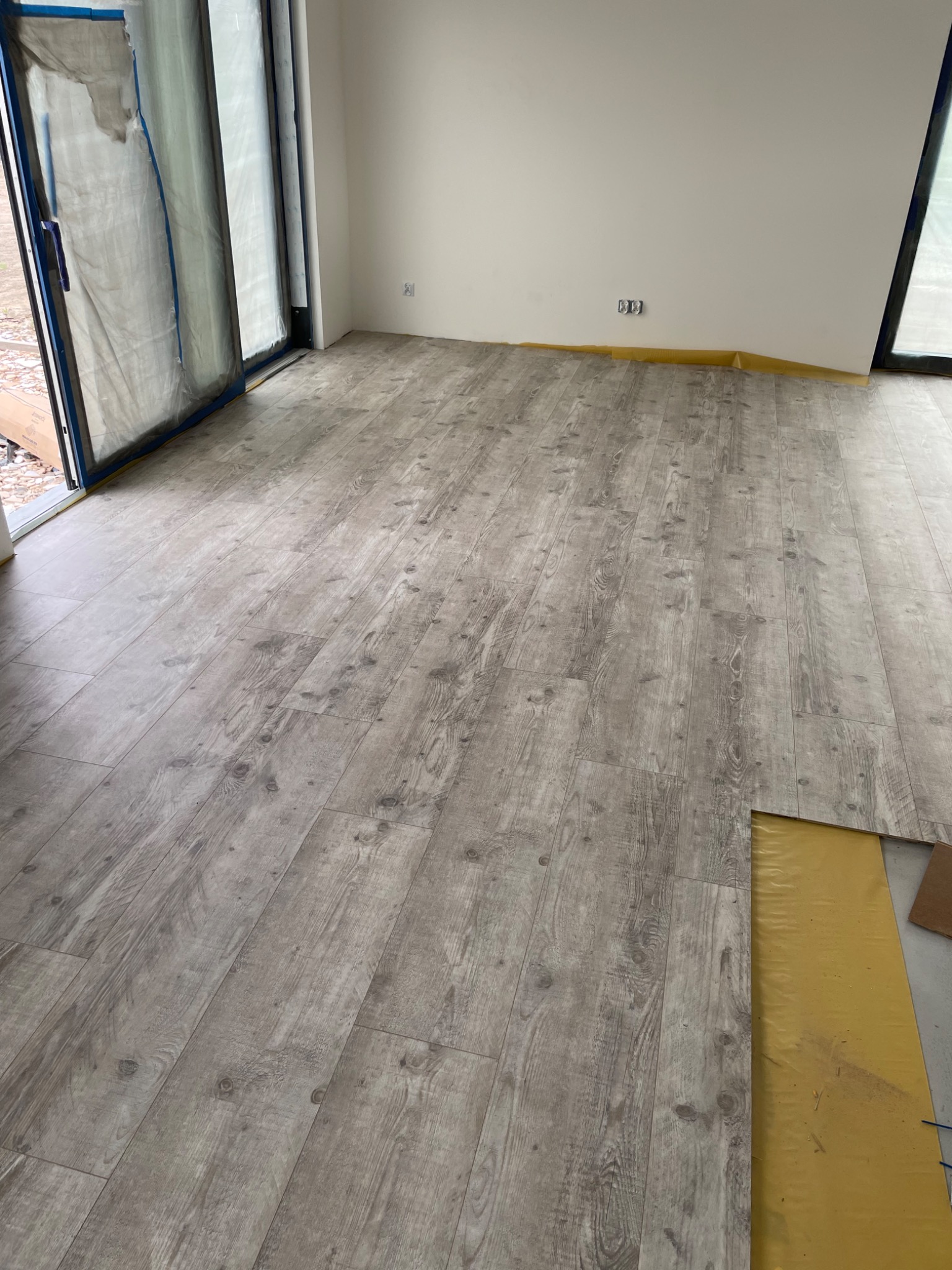 Jasnoszara podłoga laminowana z imitacją drewna, ułożona w nowym pomieszczeniu z zabezpieczonymi folią oknami i listwami przypodłogowymi zabezpieczonymi taśmą malarską, widoczny fragment podkładu.