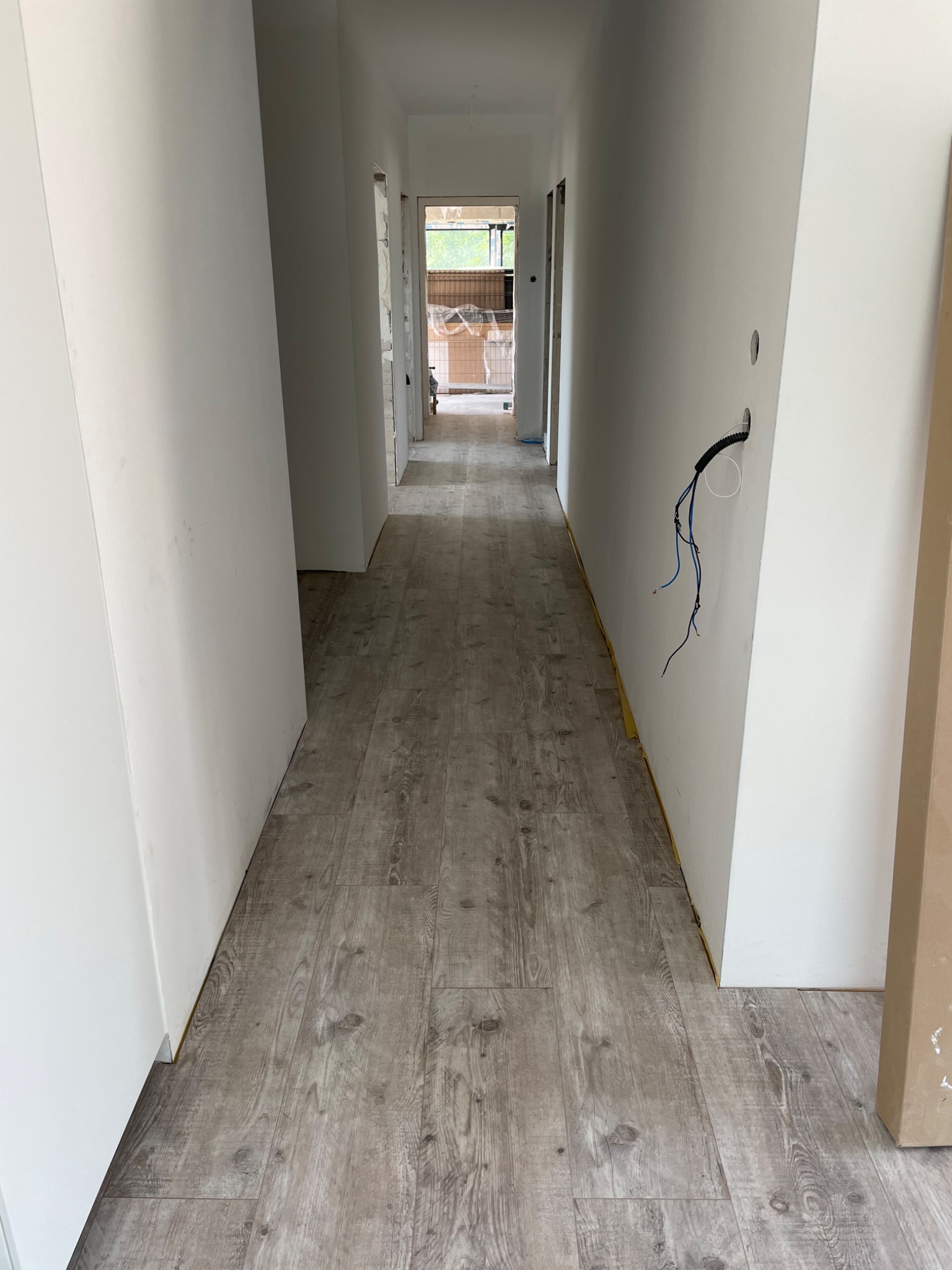 Korytarz w trakcie remontu: świeżo położona podłoga z paneli imitujących drewno, białe ściany przygotowane do malowania, wystające przewody elektryczne, widoczne zabezpieczenia podłogi przy ścianach.