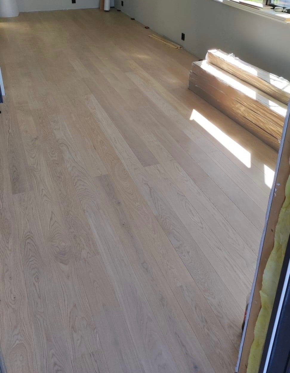 Jasny, dębowy parkiet ułożony w salonie, z prostopadle padającym światłem słonecznym i paczkami desek czekającymi na montaż przy ścianie.