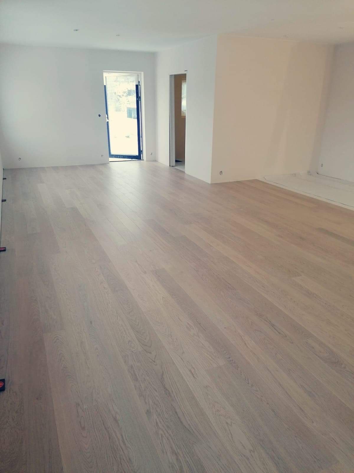 Jasny, świeżo ułożony parkiet w pustym pokoju z białymi ścianami, widoczne zabezpieczenie folią malarską przy ścianie.