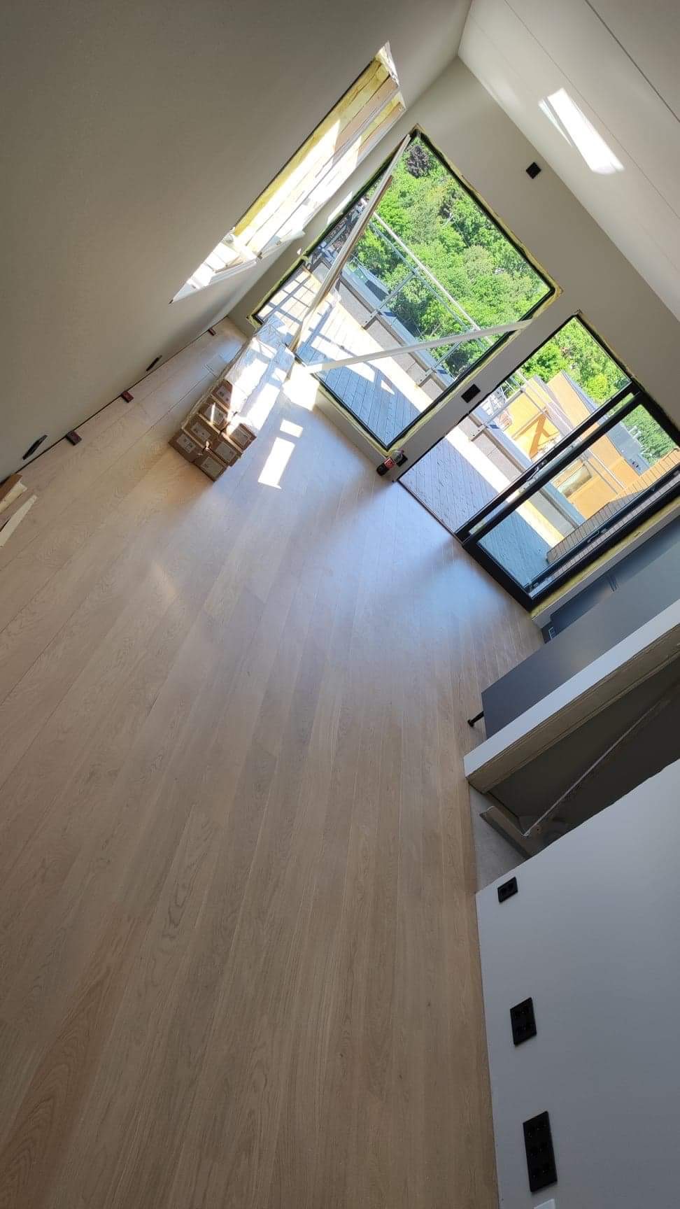 Jasny, drewniany parkiet ułożony w przestronnym salonie z dużymi oknami i widokiem na zieleń, częściowo zakryty kartonami z panelami podłogowymi.