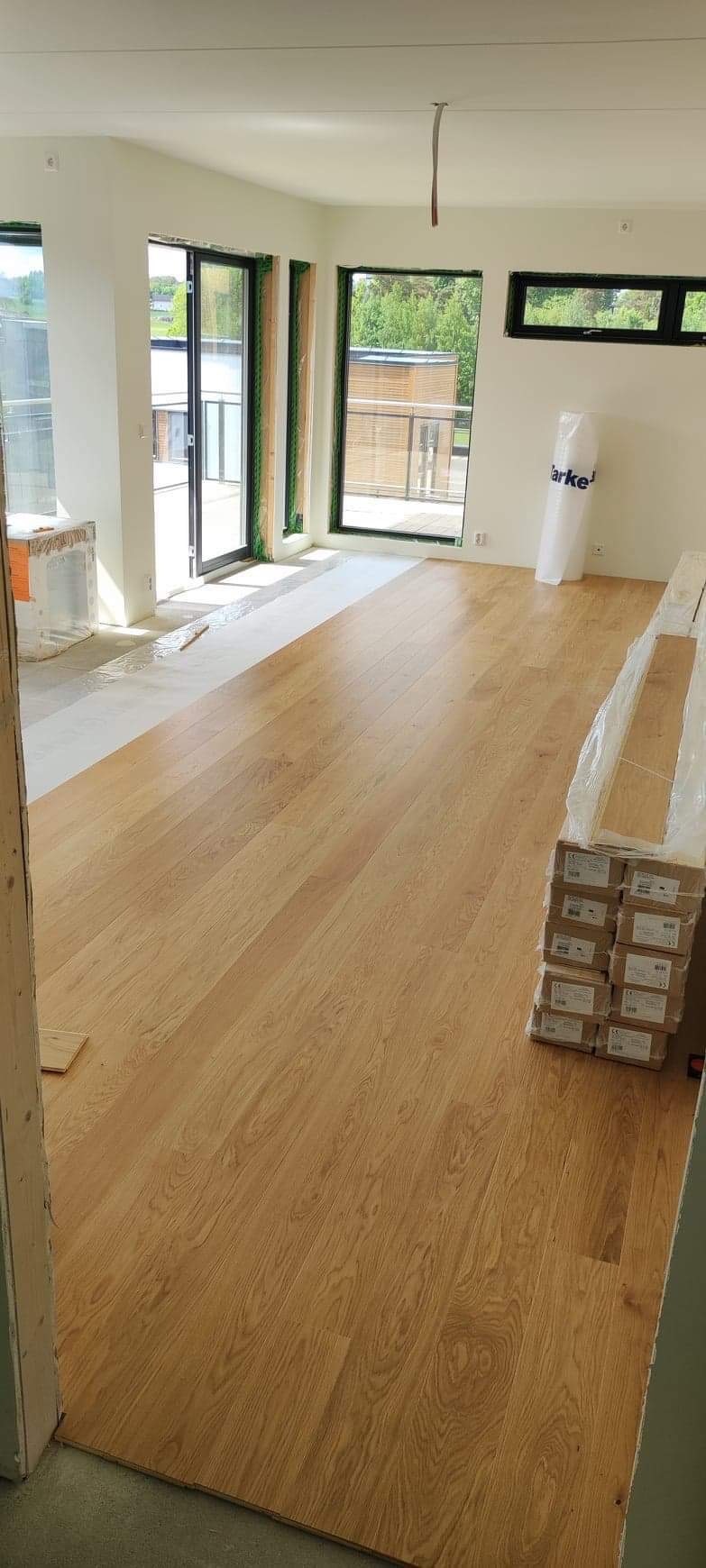 Jasny parkiet dębowy ułożony w przestronnym, słonecznym pokoju z dużymi oknami balkonowymi, widoczne kartony z panelami i folia ochronna na części podłogi.
