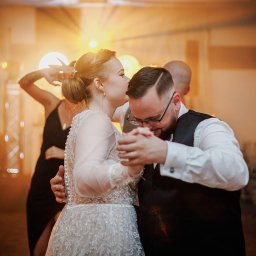 Jarosław Juraszek - Para młoda w tańcu podczas wesela, panna młoda całuje pana młodego w skroń, w tle goście i dekoracje świetlne.