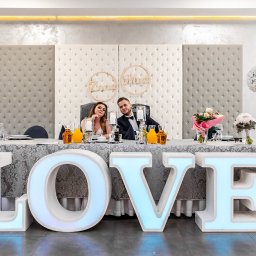 Jarosław Juraszek - Para młoda siedząca za stołem udekorowanym kwiatami i napisem LOVE z podświetlanych liter, za nimi dekoracyjne panele tapicerowane.