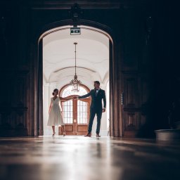 Jarosław Juraszek - Para młoda trzymająca się za ręce w eleganckim, ciemnym holu z drewnianą podłogą i dużymi drzwiami w tle, oświetlonymi światłem z zewnątrz.