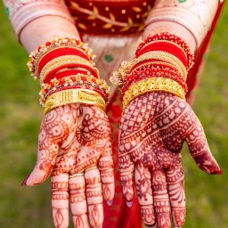 Jarosław Juraszek - Dłonie kobiety zdobione henną i licznymi, kolorowymi bransoletami, prezentowane na tle czerwonej, wzorzystej sukni.