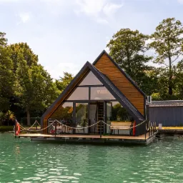 Dom w kształcie litery A na wodzie, z drewnianą elewacją i dużymi oknami, otoczony zielenią drzew, z pomostem i linami zabezpieczającymi.