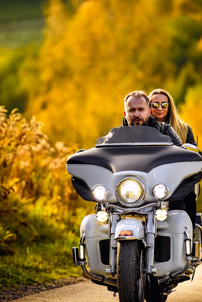Para na motocyklu Harley Davidson Electra Glide, złote światło jesiennego popołudnia, rozmyte tło z drzewami w odcieniach żółci i zieleni.
