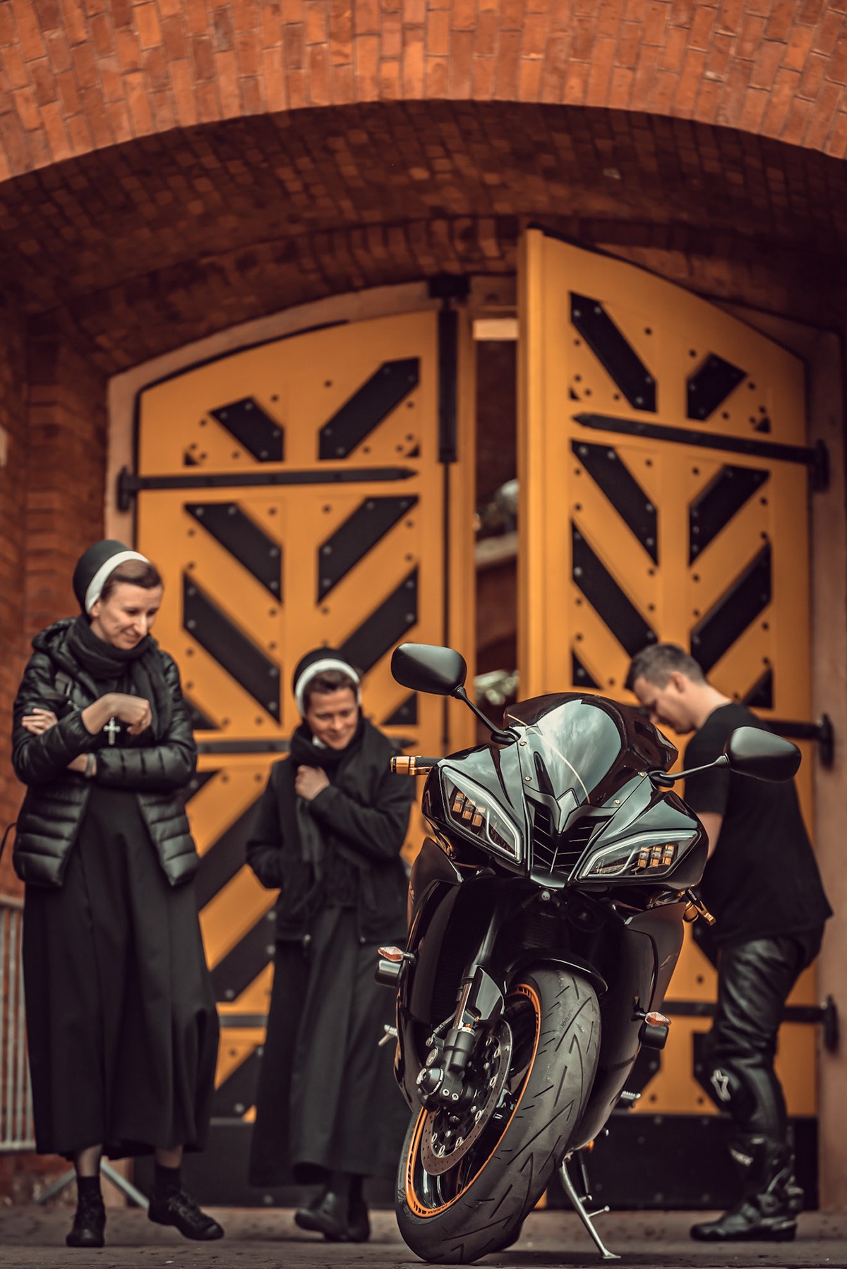Dwie zakonnice i mężczyzna w czarnych strojach pozują z czarnym motocyklem sportowym przed otwartymi, żółtymi drzwiami z czarnymi wzorami w kształcie strzałek.