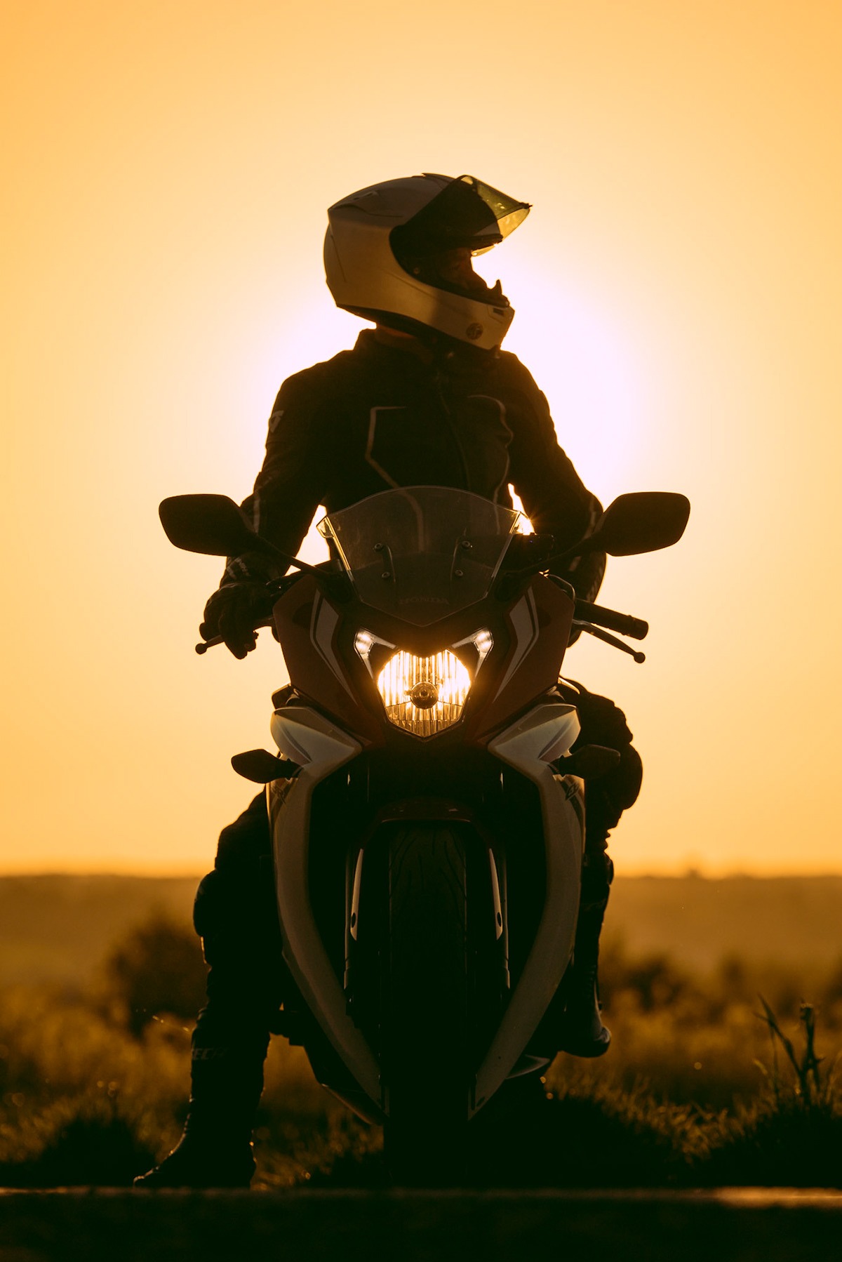 Sylwetka motocyklisty w kasku na motocyklu Honda na tle zachodzącego słońca, z włączonymi światłami przednimi.