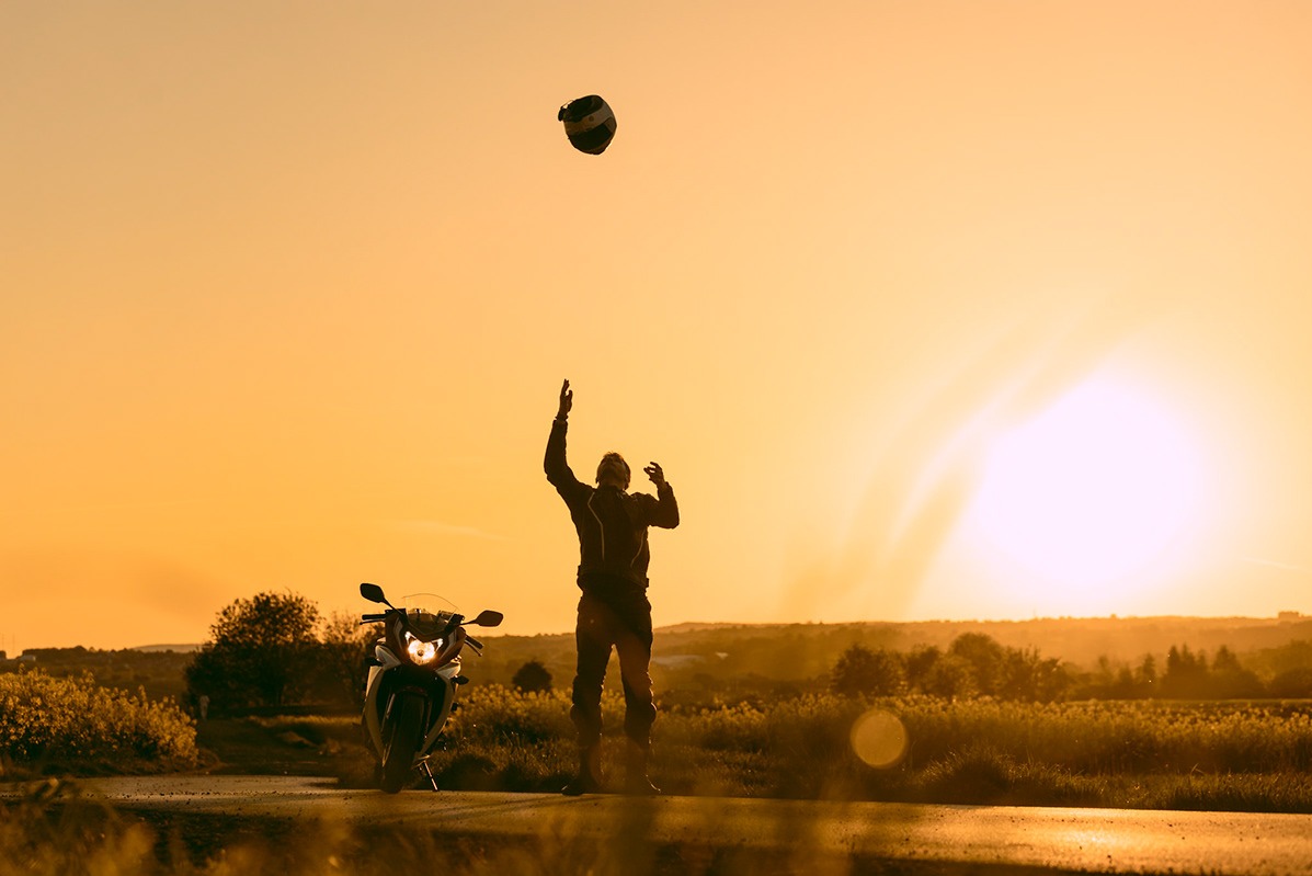 Sylwetka motocyklisty rzucającego kaskiem w górę, motocykl zaparkowany obok, ciepłe światło zachodzącego słońca w tle.