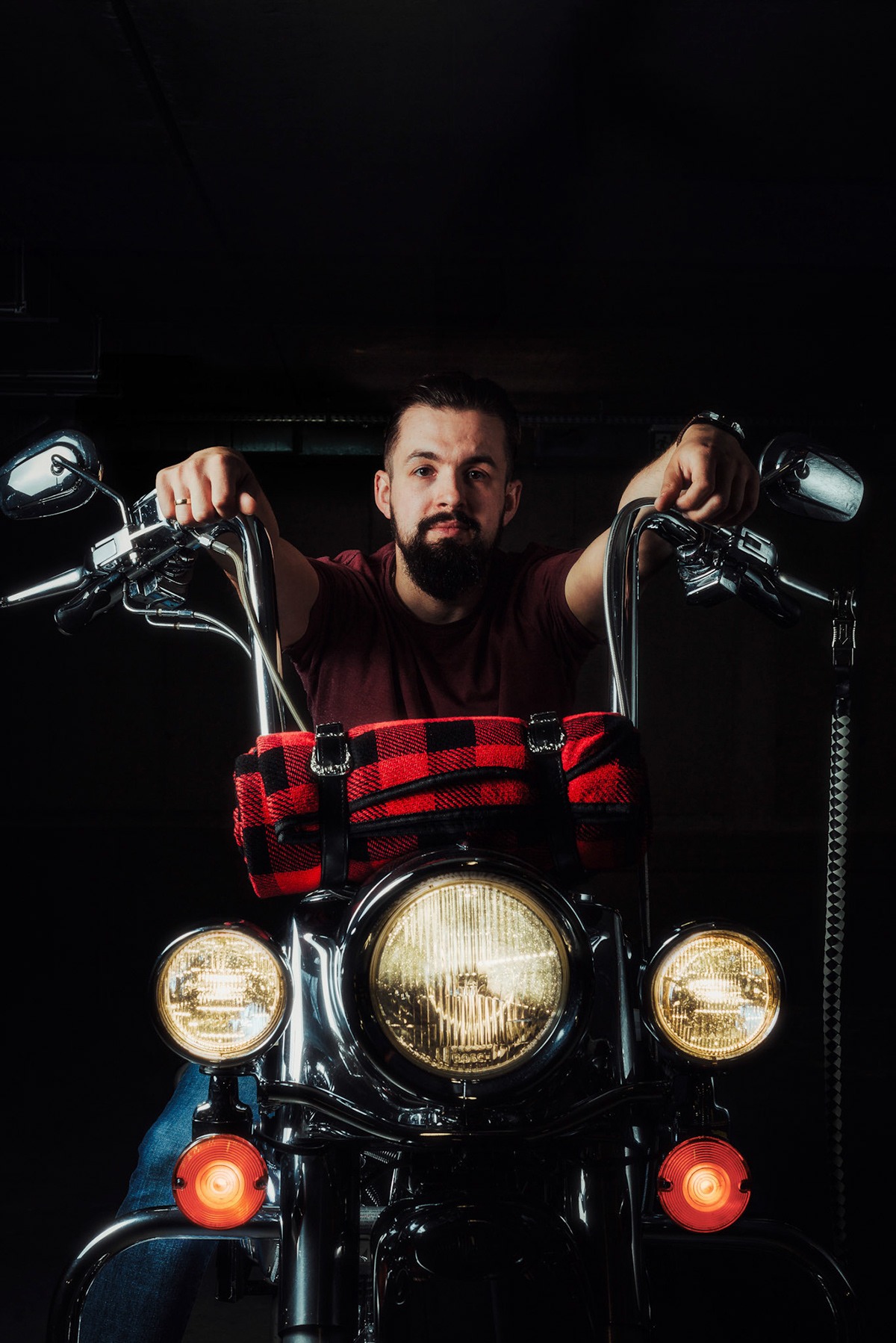 Mężczyzna z brodą opiera ręce na kierownicy motocykla z włączonymi światłami, z przodu torba w czerwoną kratę.