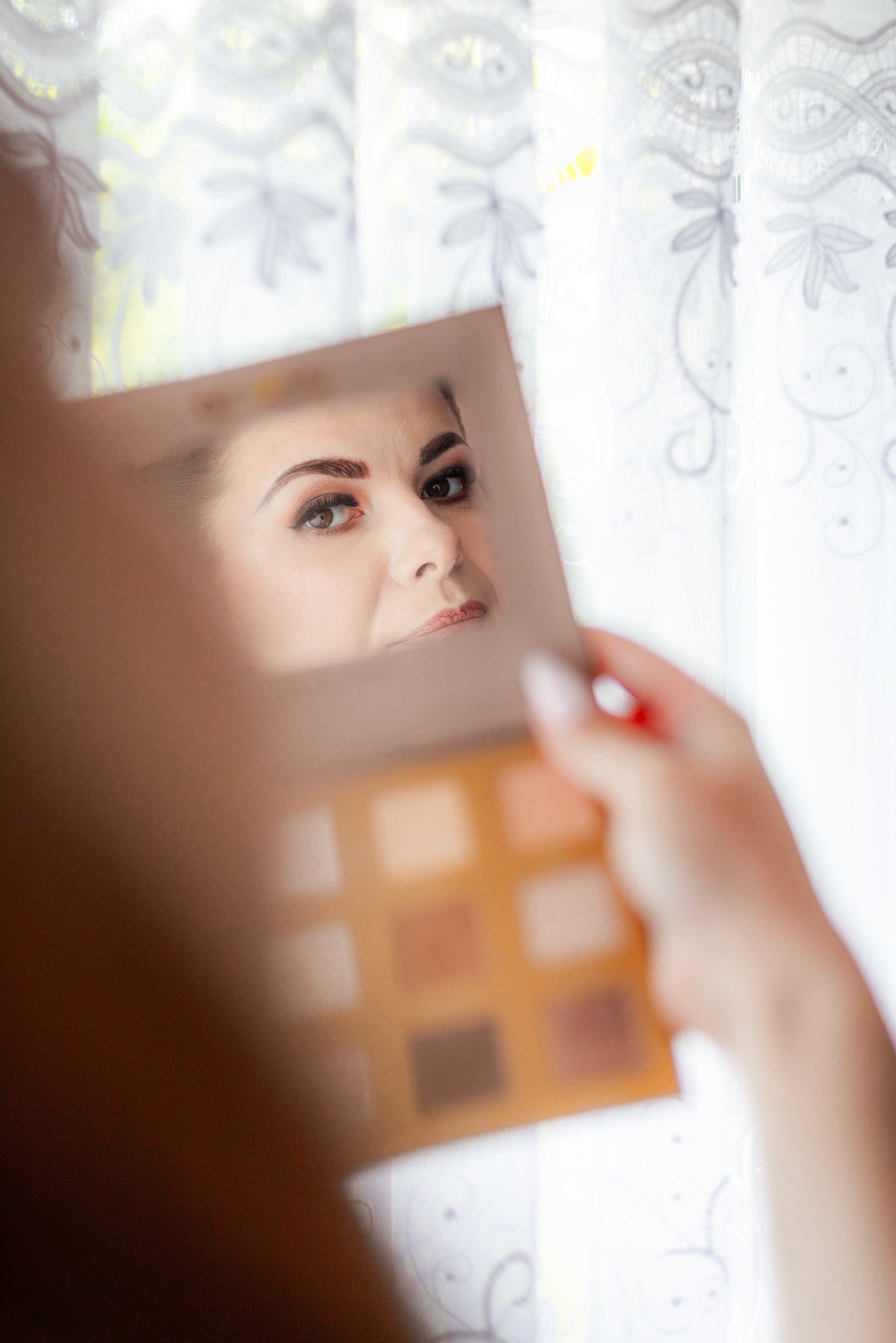 Kobieta przegląda się w lusterku, trzymając paletę cieni do powiek, widoczna twarz z wyraźnie podkreślonym makijażem oczu na tle firanki.