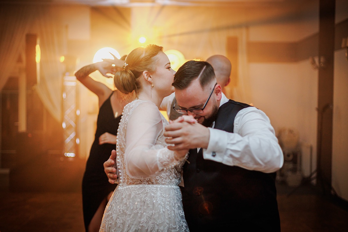 Para młoda w tańcu podczas wesela, panna młoda całuje pana młodego w skroń, w tle goście i dekoracje świetlne.