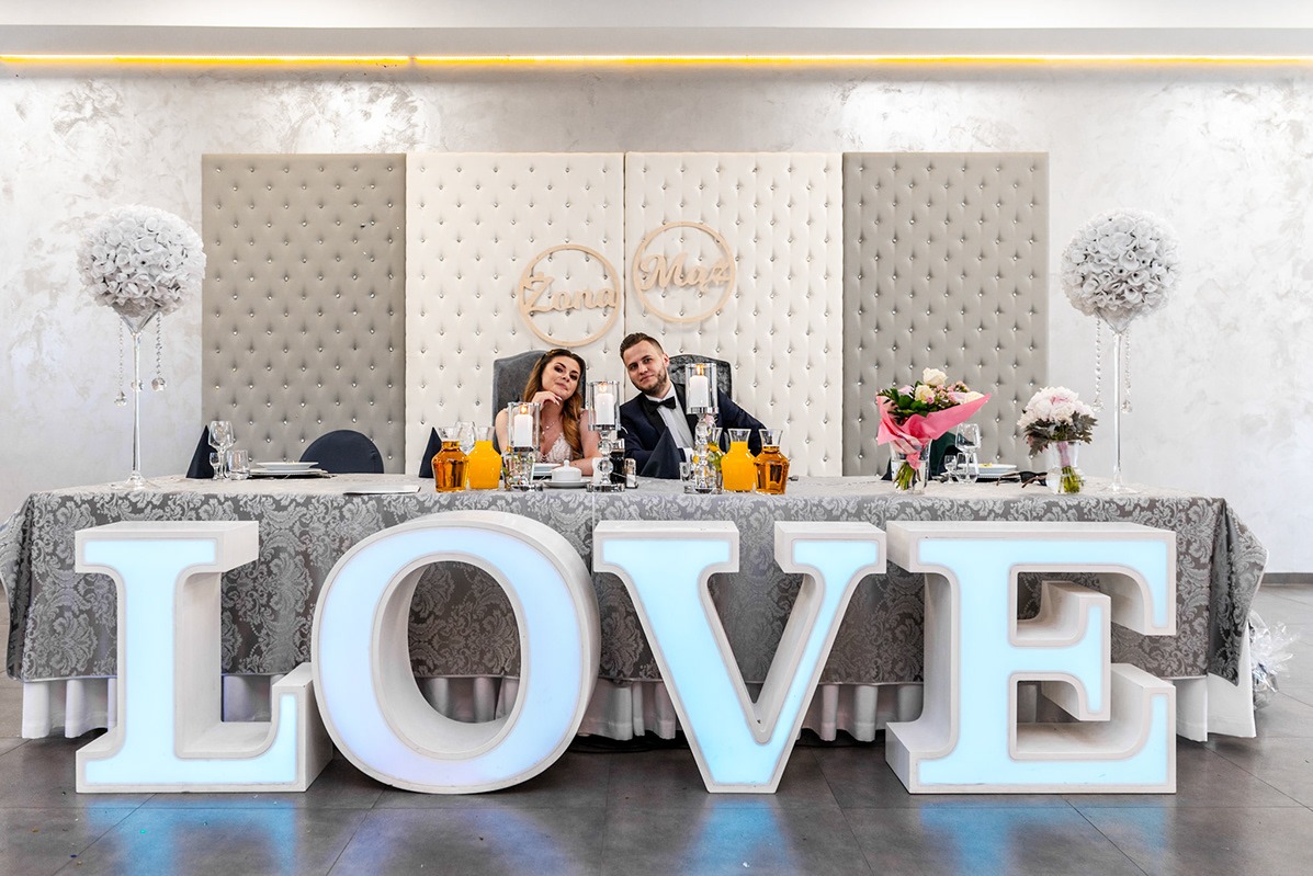 Para młoda siedząca za stołem udekorowanym kwiatami i napisem LOVE z podświetlanych liter, za nimi dekoracyjne panele tapicerowane.