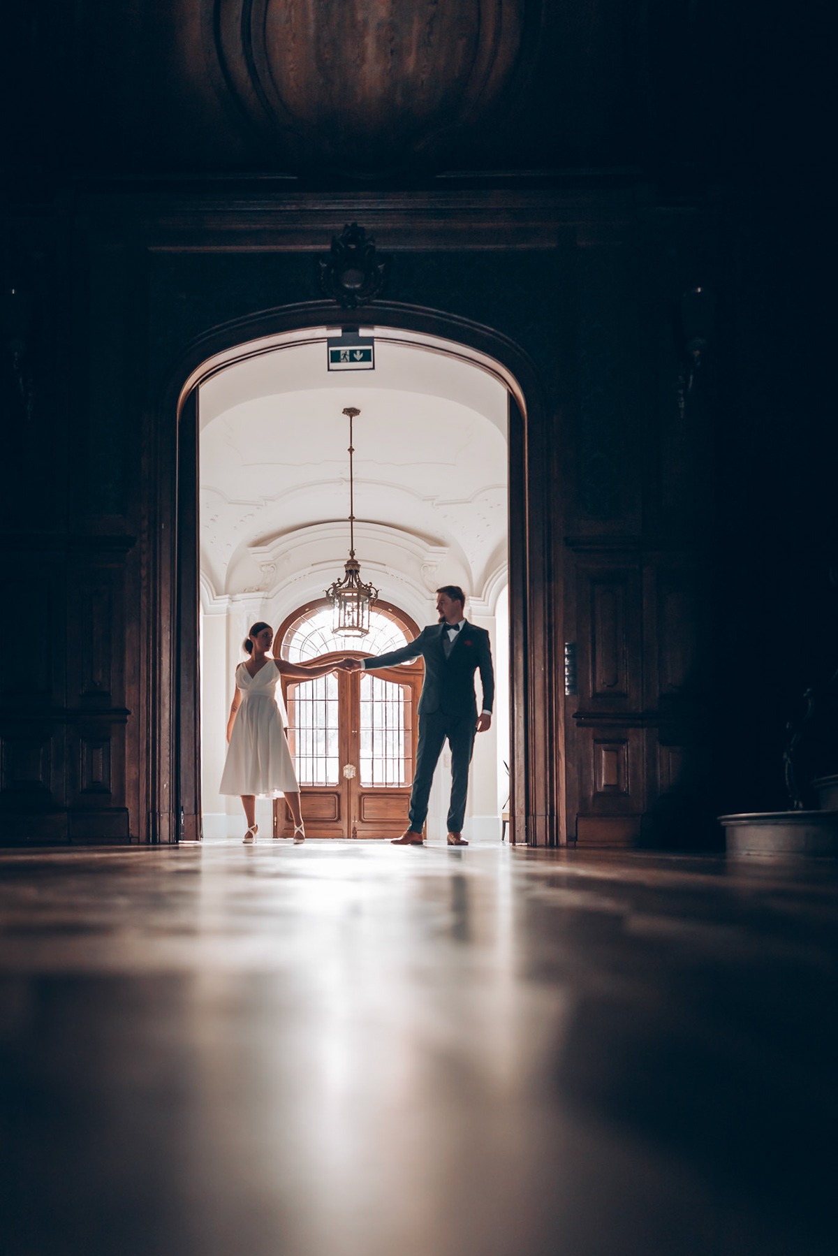 Para młoda trzymająca się za ręce w eleganckim, ciemnym holu z drewnianą podłogą i dużymi drzwiami w tle, oświetlonymi światłem z zewnątrz.