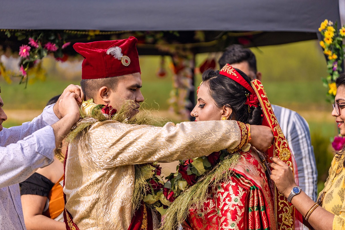 Para młoda w tradycyjnych strojach ślubnych podczas ceremonii, mężczyzna w czerwonym kapeluszu ozdobionym piórem, kobieta w czerwonym welonie z haftowanymi złotymi kwiatami, oboje z girlandami...