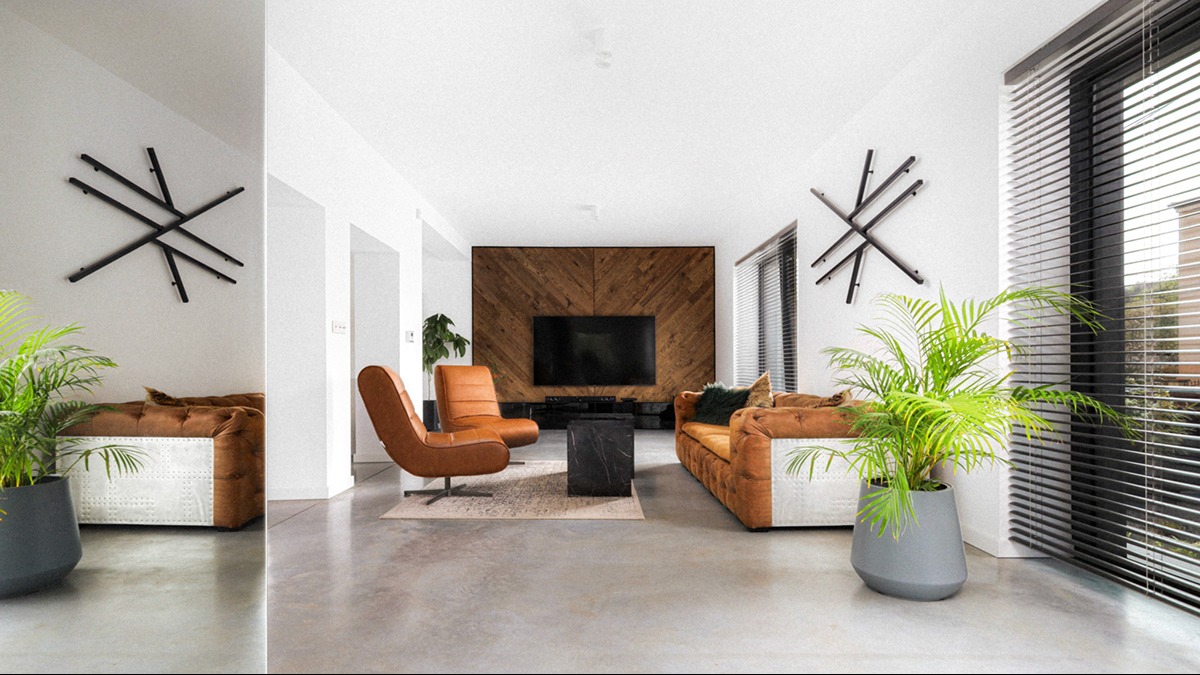 Jasne, przestronne wnętrze salonu z brązową sofą, skórzanymi fotelami, drewnianą ścianą z telewizorem, nowoczesnymi dekoracjami ściennymi i roślinami doniczkowymi w szarych donicach.