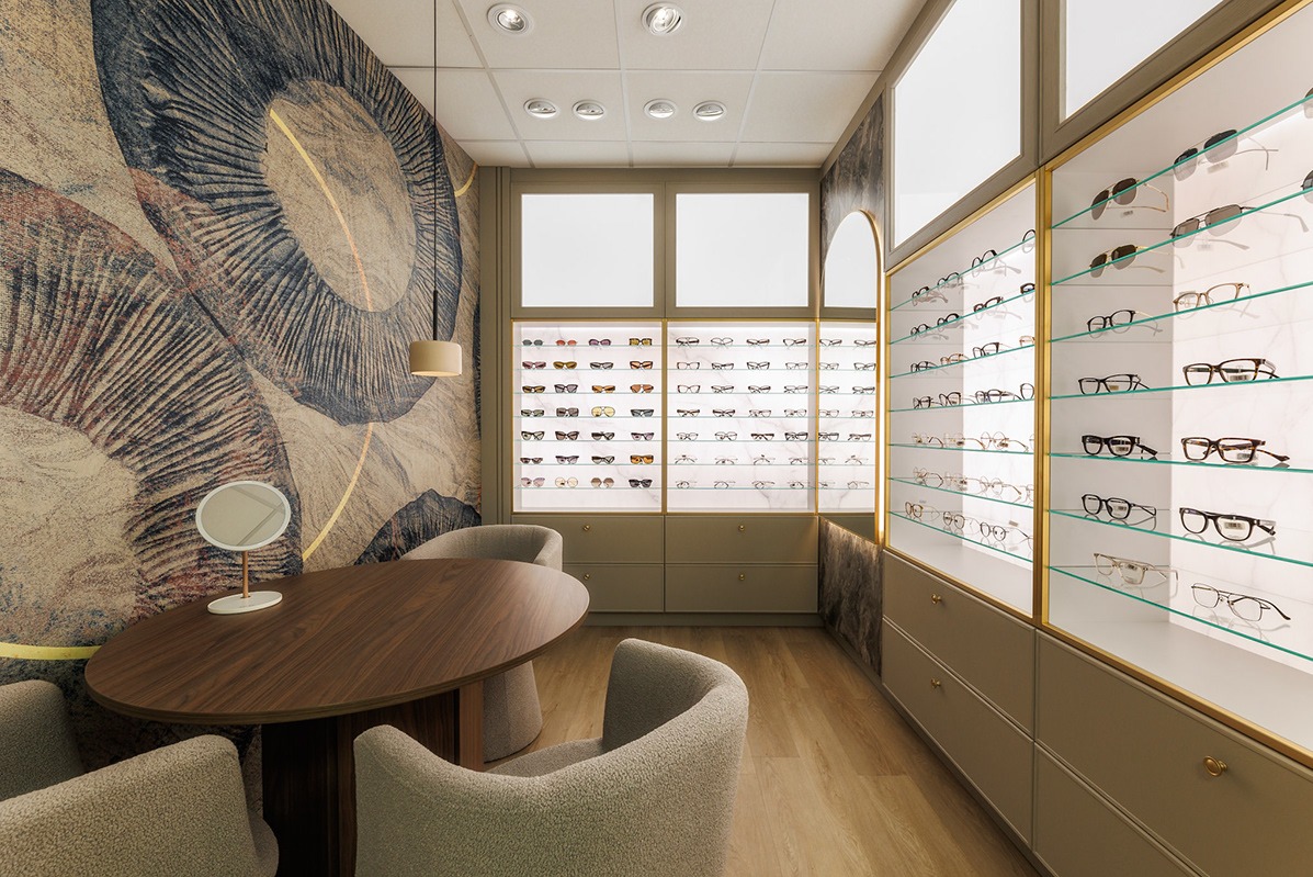 Wnętrze salonu optycznego z ekspozycją okularów w podświetlanych gablotach, drewnianym stołem i tapetą z motywem abstrakcyjnych kwiatów.