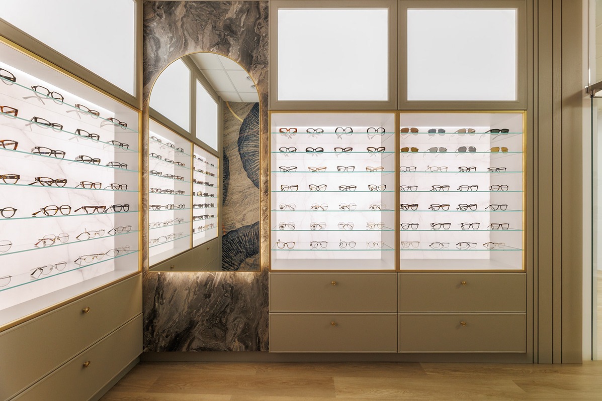 Wnętrze salonu optycznego z ekspozycją okularów na podświetlanych półkach, marmurowa ściana z lustrem w kształcie łuku, beżowe szafki z okrągłymi uchwytami, drewniana podłoga.
