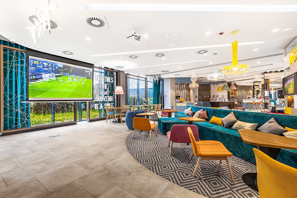 Nowoczesne wnętrze hotelowe z dużym ekranem wyświetlającym mecz piłki nożnej, turkusową sofą z poduszkami, różowymi i pomarańczowymi krzesłami oraz geometrycznym dywanem.