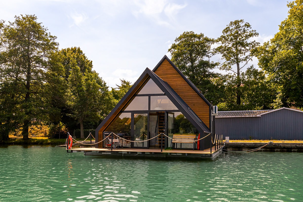 Dom w kształcie litery A na wodzie, z drewnianą elewacją i dużymi oknami, otoczony zielenią drzew, z pomostem i linami zabezpieczającymi.