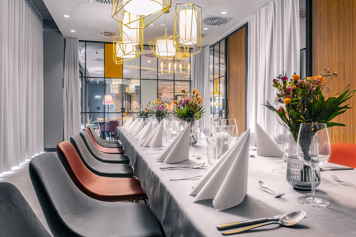 Uroczysty stół w restauracji, nakryty białym obrusem, z rzędami szarych i ceglastych krzeseł, białymi serwetkami ułożonymi w trójkąty, szklanymi naczyniami, sztućcami ze złotymi akcentami...