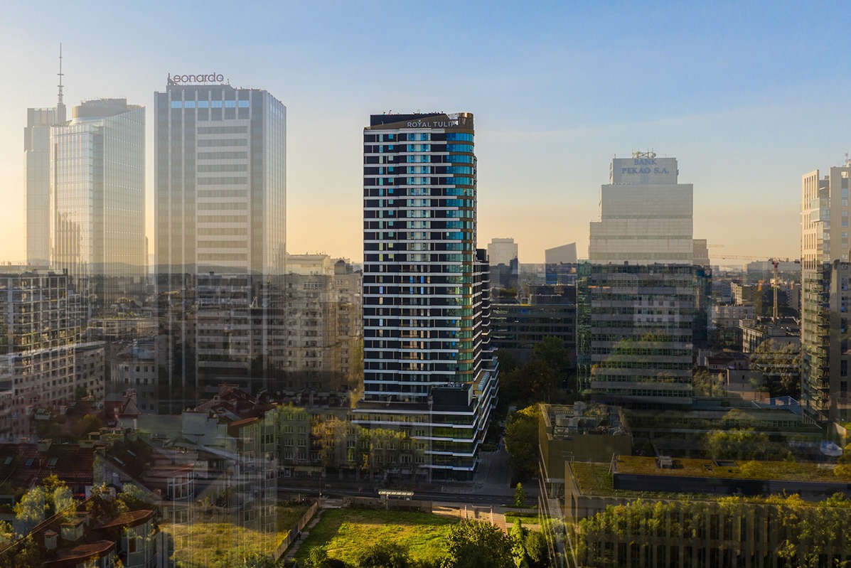 Panorama Warszawy o wschodzie słońca z widokiem na nowoczesne wieżowce, w tym Royal Tulip, Leonardo i Pekao S.A., zieleń miejska w dolnej części kadru.