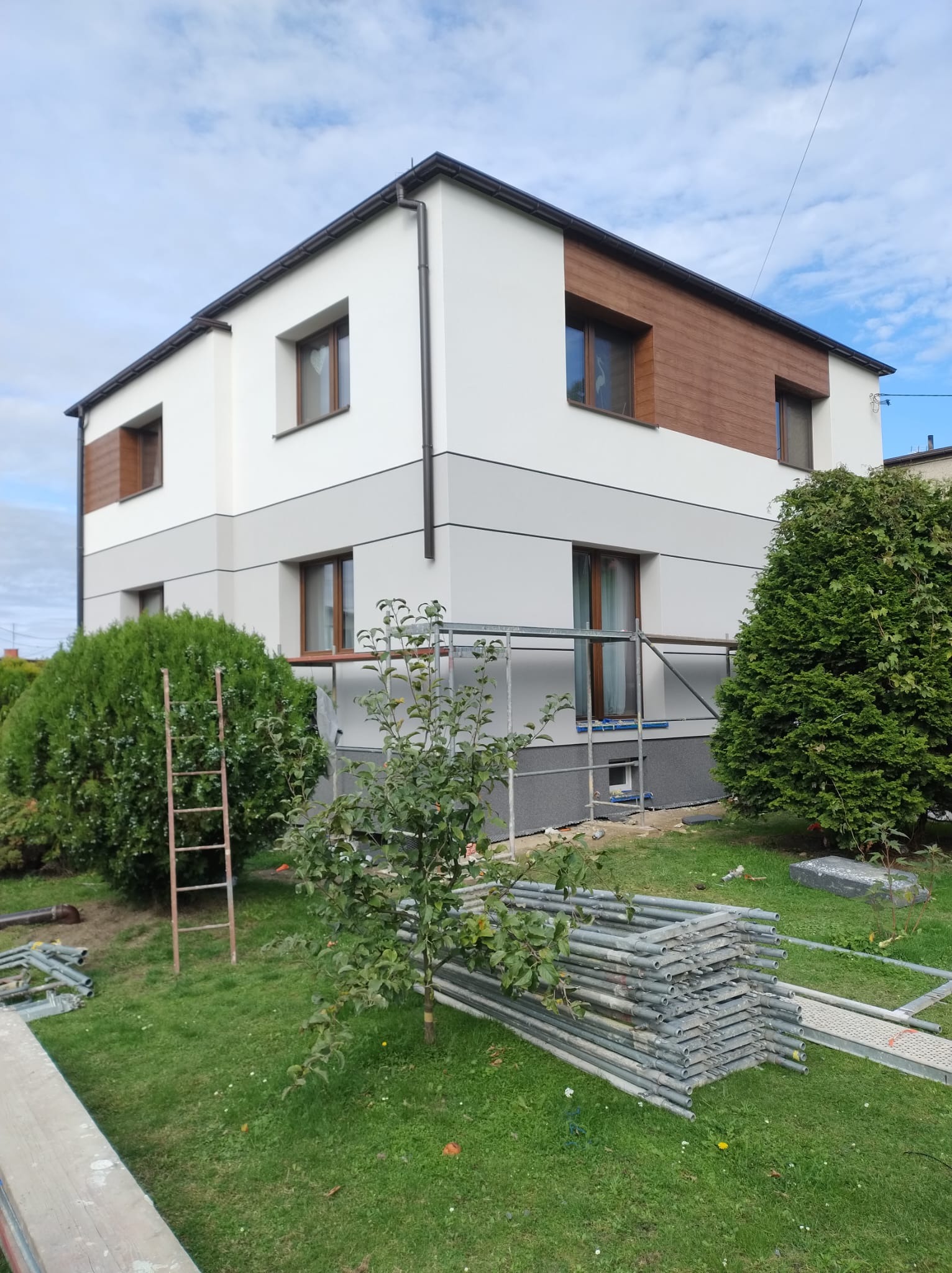 Dwukondygnacyjny dom po renowacji elewacji, z elementami drewnianymi i rusztowaniem. Widoczne detale wykończenia i otoczenie budynku.