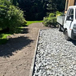 Przygotowanie terenu pod ścieżkę: wyrównana ziemia obok utwardzonej powierzchni z szarym kruszywem, ograniczona betonową obwódką, z widocznym białym samochodem dostawczym i zielenią w tle.