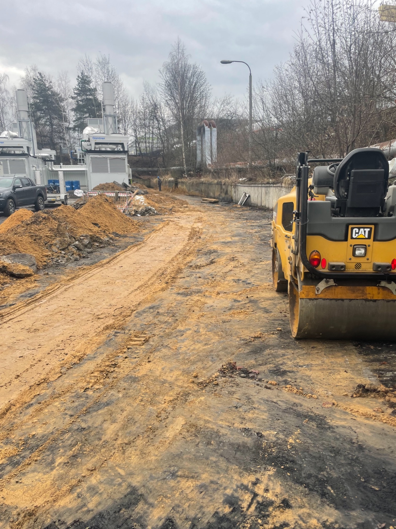 Żółty walec drogowy CAT na placu budowy, częściowo utwardzona droga z piasku i ziemi, w tle instalacje przemysłowe i zaparkowany samochód, pochmurne niebo.