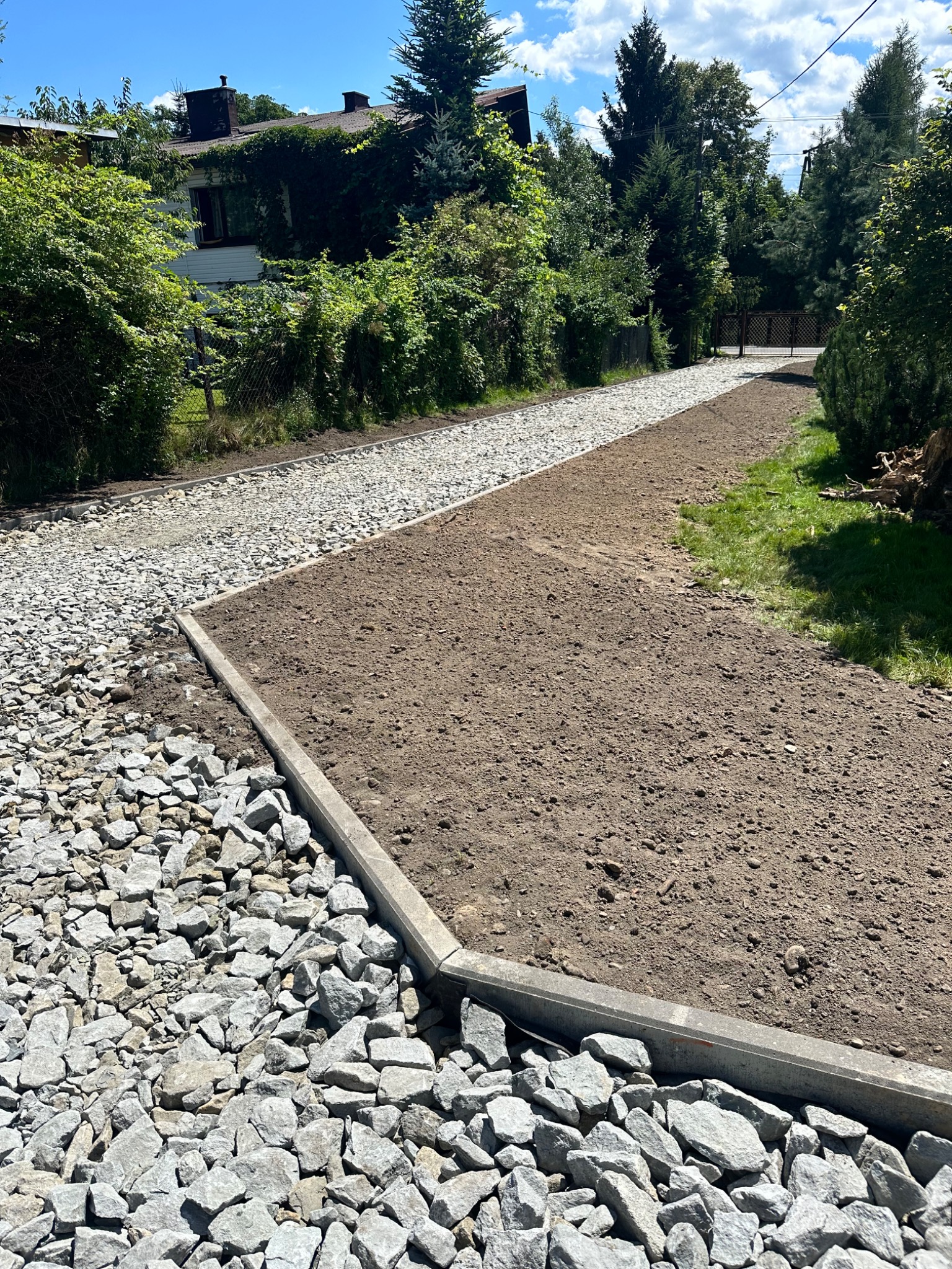 Przygotowany teren pod budowę drogi dojazdowej: wytyczone krawężniki, wysypany kamień i wyrównana ziemia, widok z poziomu gruntu.