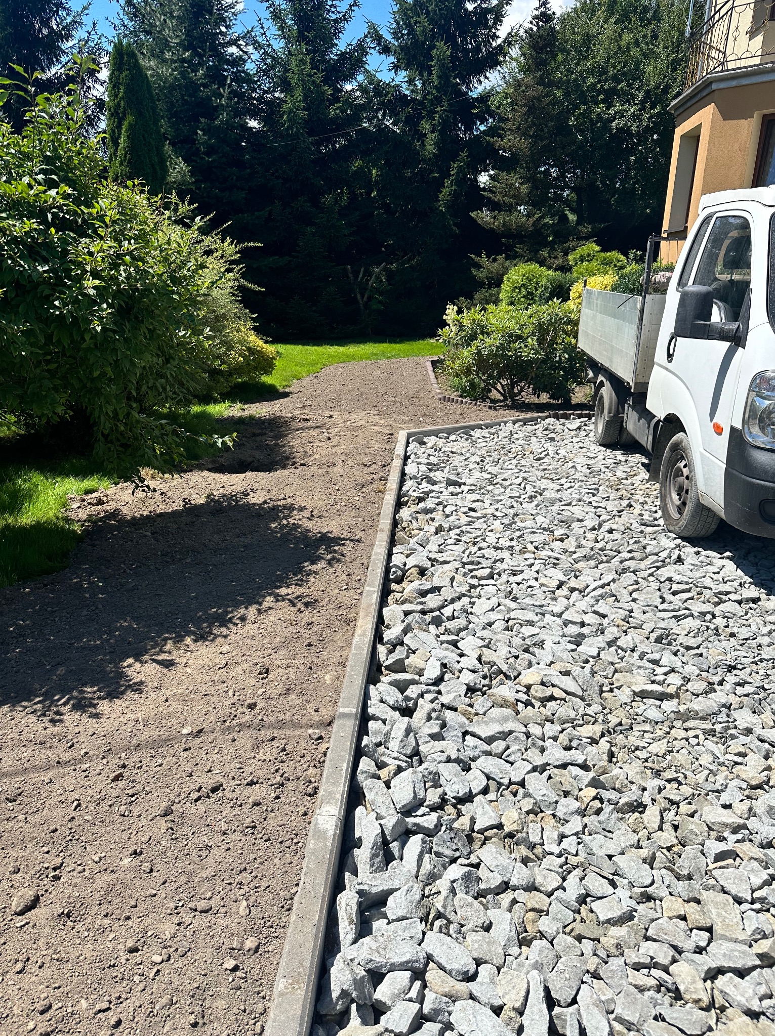 Przygotowanie terenu pod ścieżkę: wyrównana ziemia obok utwardzonej powierzchni z szarym kruszywem, ograniczona betonową obwódką, z widocznym białym samochodem dostawczym i zielenią w tle.