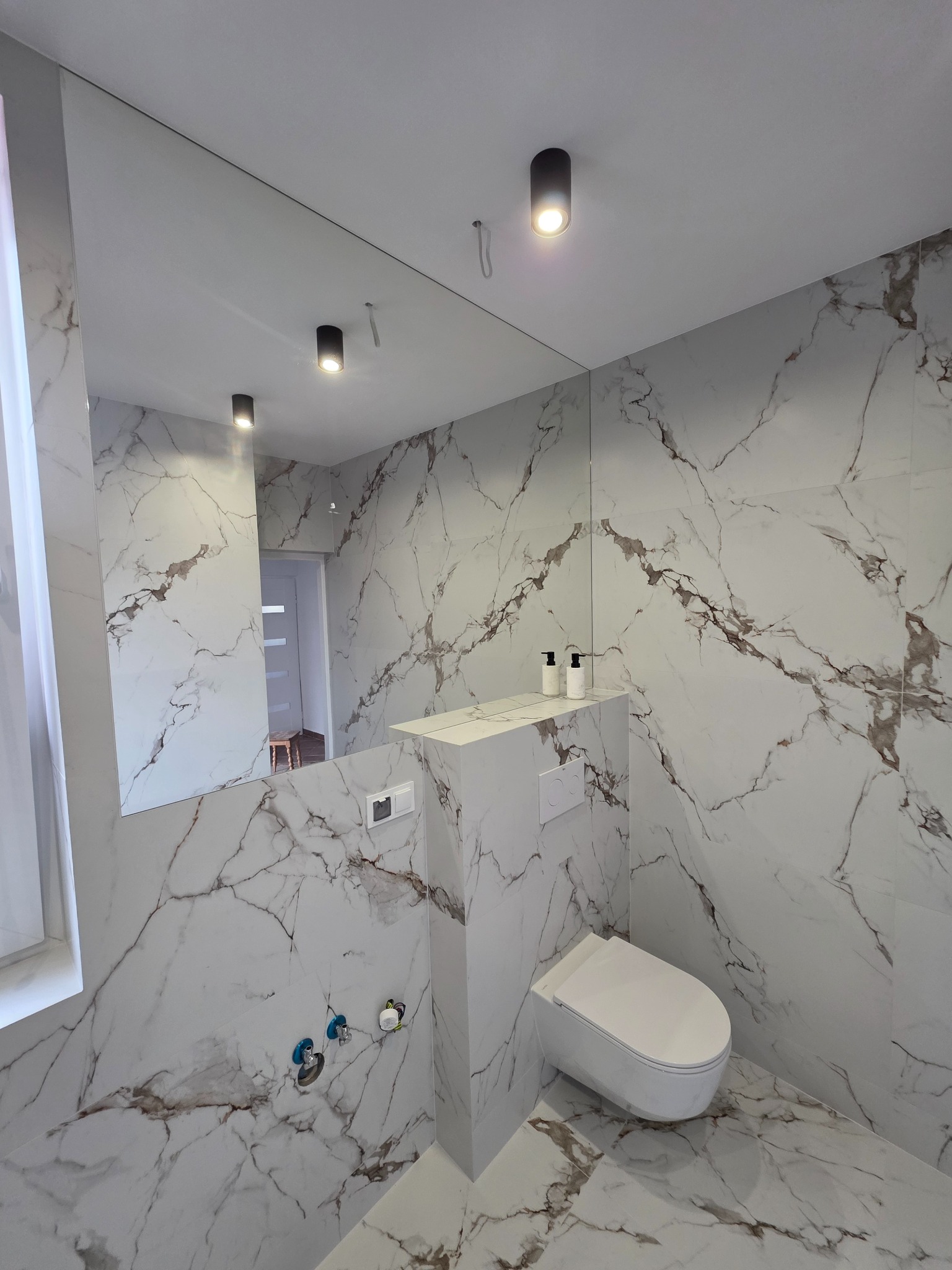 Elegancka łazienka z marmurowymi płytkami, podwieszaną toaletą i dużym lustrem. Widoczne czarne lampy sufitowe i minimalistyczne akcesoria. Nowoczesny design.