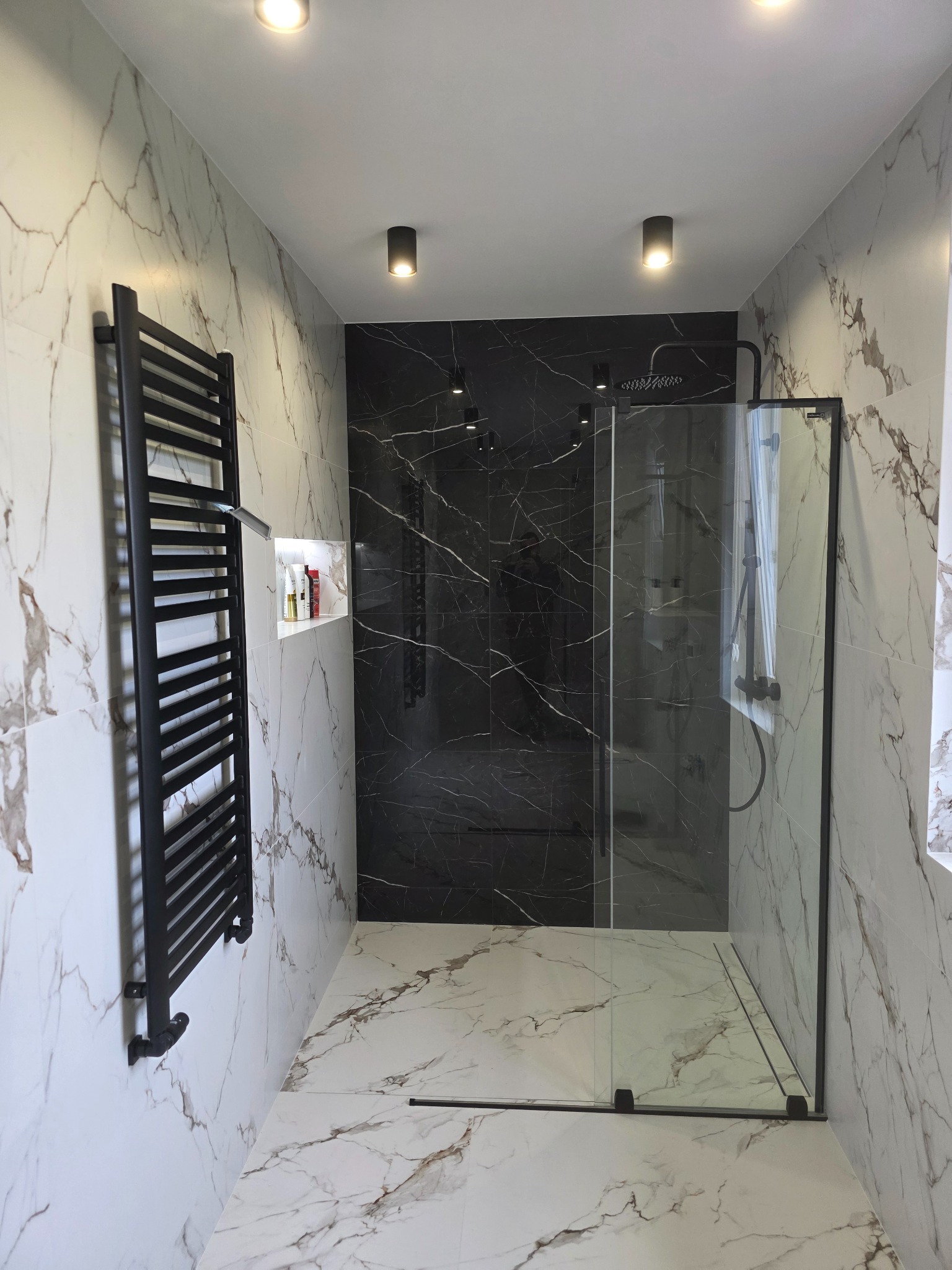 Nowoczesna łazienka z kabiną typu walk-in, czarną armaturą i grzejnikiem drabinkowym na tle płytek imitujących marmur. Minimalistyczny design, oświetlenie punktowe.