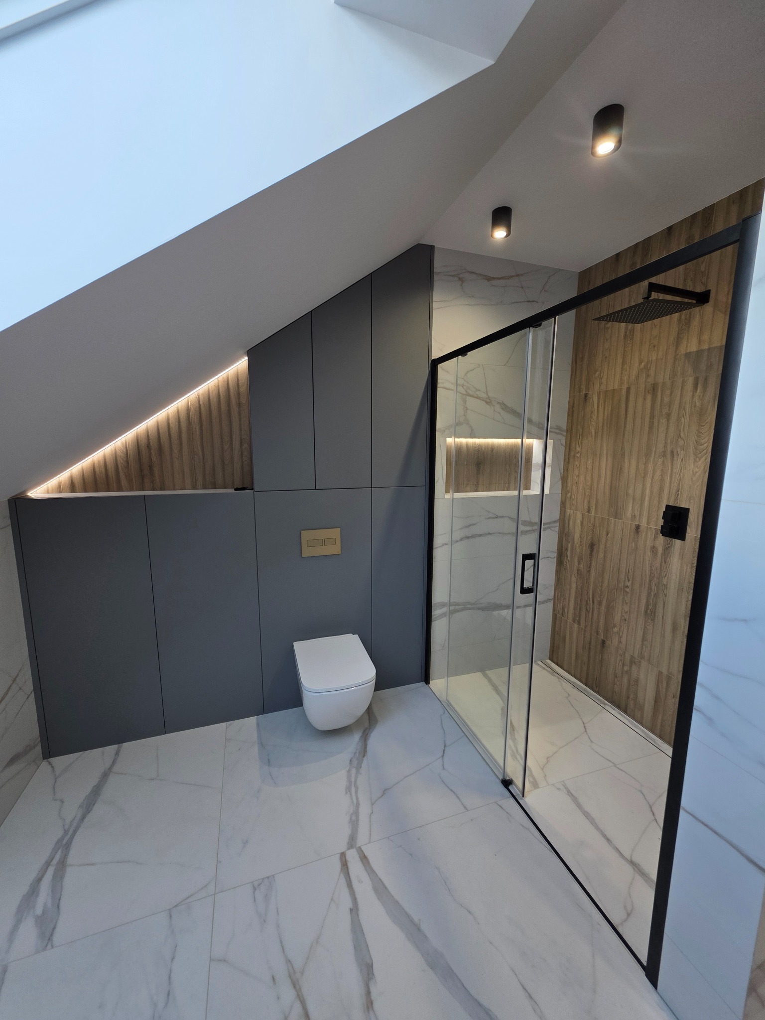 Nowoczesna łazienka na poddaszu z kabiną prysznicową typu walk-in, podświetlanymi wnękami i marmurową podłogą. Uwagę zwraca minimalistyczna toaleta i szara zabudowa.