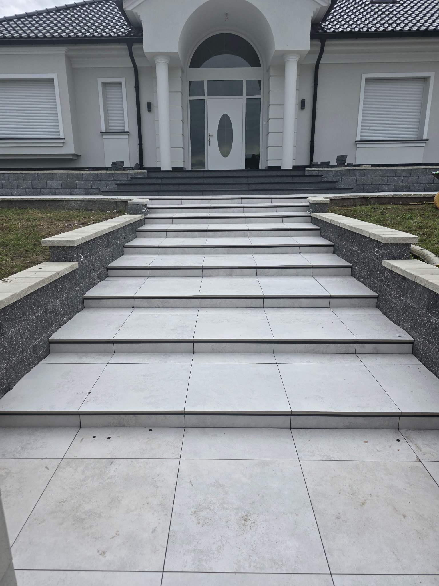 Białe płytki na schodach wejściowych do domu z kolumnami, widok z dołu. Szare obrzeża i kostka. Nowoczesny design.