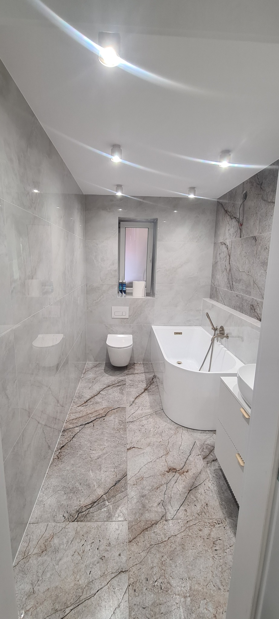 Nowoczesna łazienka z marmurowymi płytkami, białą wanną i umywalką. Minimalistyczny design, oświetlenie punktowe. Widok na toaletę i okno w tle.