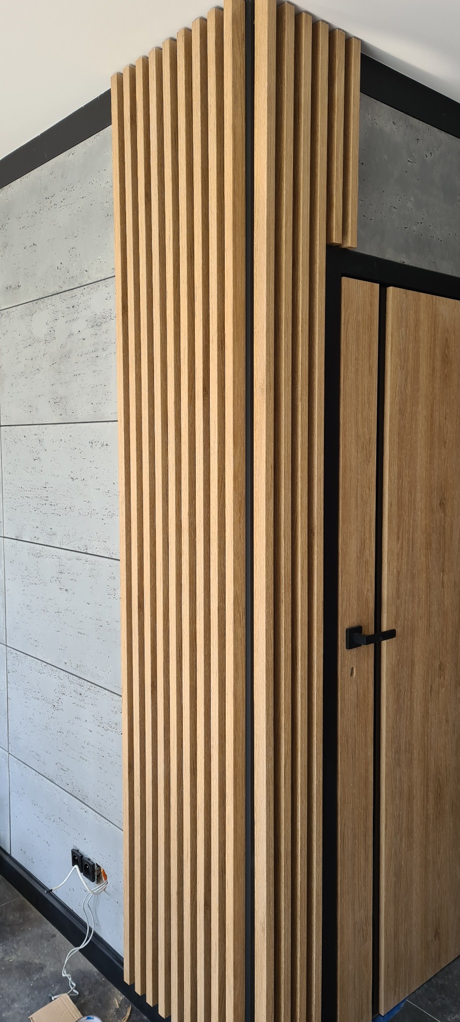 Drewniane lamele na ścianie, betonowe płytki, czarna listwa przypodłogowa i fragment drzwi z czarną ościeżnicą. Widoczne gniazdko elektryczne.