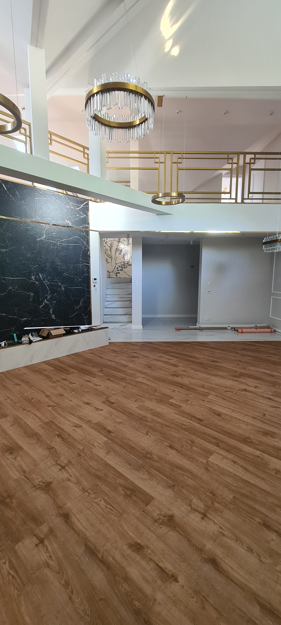 Nowoczesne wnętrze z drewnianą podłogą, marmurową ścianą i designerskimi lampami. Widoczne schody i balustrada z metalowymi zdobieniami. Jasne, przestronne pomieszczenie.